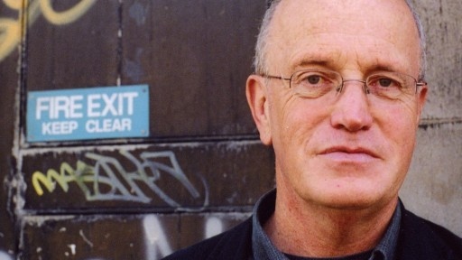 Cult author and psychogeographer Iain Sinclair infront of an old, grafffitied wall.