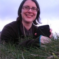 A picture of MLF Children and Young People's Coordinator Liz Postlethwaite sitting in a field, smiling, wearing headphones