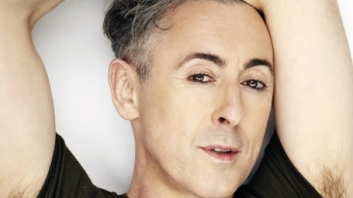 Image of Alan Cumming with both arms crossed above his head, and his armpit hair showing above his t-shirt sleeves