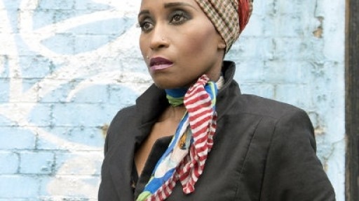 Image of Malika Booker in a brown and orange checked head scarf, standing in front of a pale blue, grafitied wall