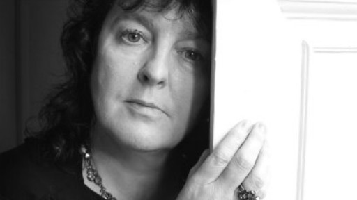 Black and white head and shoulders shot of Carol Ann Duffy, holding on to a white door whilst looking straight ahead