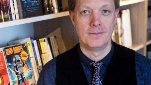 Head and shoulders shot of Adam Roberts wearing shirt, tie and waistcoat, standing next to bookshelves, including his own book Adam Robots