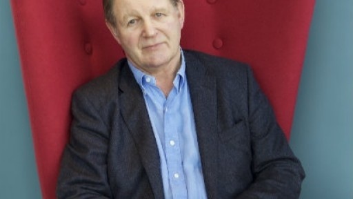 Image of Michael Morpurgo, sitting in a wingback red velvet chair, with his hands on his leg, smiling