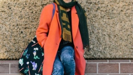 Photo of author Olivia Laing, wearing a bright orange coat and sunglasses and leaning on a wall with one foot resting on the wall