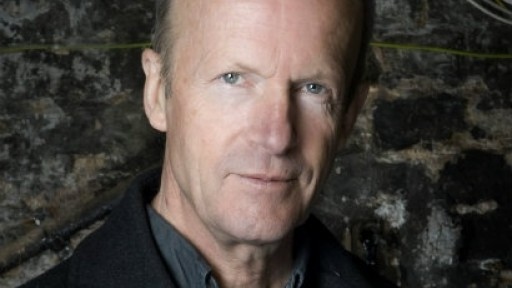 Head shot of author Jim Crace, against a dark stone walll