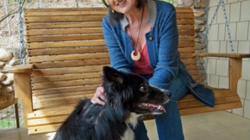 Photo of author Barbara Kingsolver, seated in a chair on a porch, stroking a black and white dog