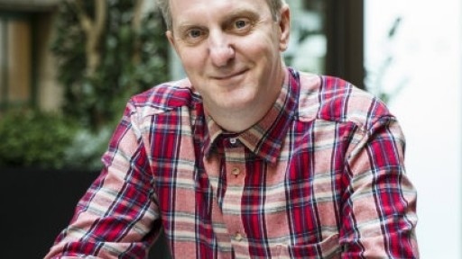 Head and shoulders shot of poet Tony Walsh, wearing a red checked shirt, sitting outside, with his hands interlaced on a table