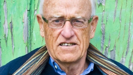 Poet CK Stead wearing a blue jumper and scarf, infront of a vivid green wall.