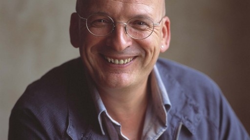Cult Irish author Roddy Doyle grinning at the camera, dressed in a blue shirt and jacket. .