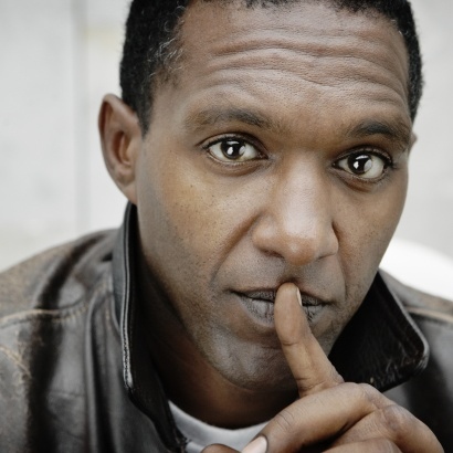 Close-up portrait of Mancunian poet Lemn Sissay in a leather jacket with his finger pressed to his lips.