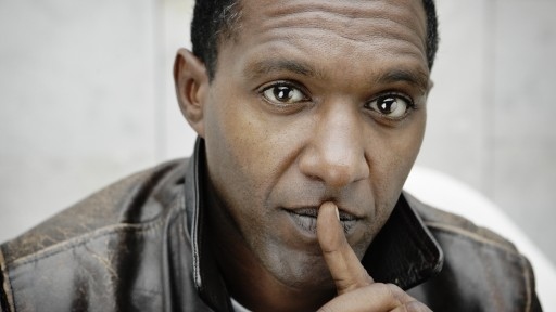 Close-up portrait of Mancunian poet Lemn Sissay in a leather jacket with his finger pressed to his lips.