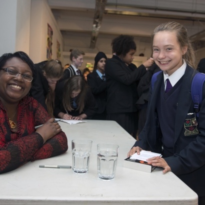 A picture of Malorie Blackman sitting at a table during a book signing talking to a young fan